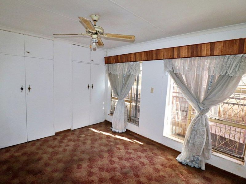 A vacant bedroom featuring built-in cupboards, a ceiling fan, and security bars on the windows, with dated carpet flooring throughout.