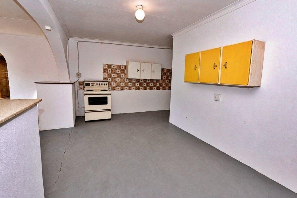 A dated, empty kitchen area featuring a freestanding stove, wall-mounted cabinets, and a tiled backsplash, requiring modernization and floor repairs.