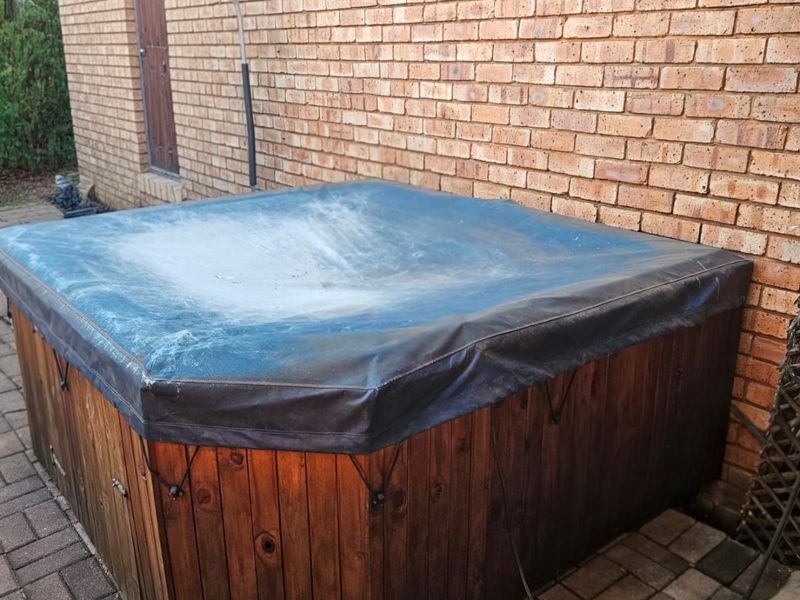 A rear exterior area featuring a brick wall and a covered hot tub set on a paved patio surface.