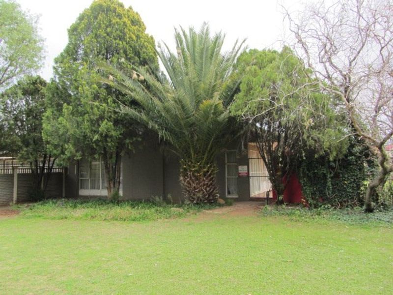 Front view of the property featuring a large palm tree, established garden, paved walkway, and security gate entrance.