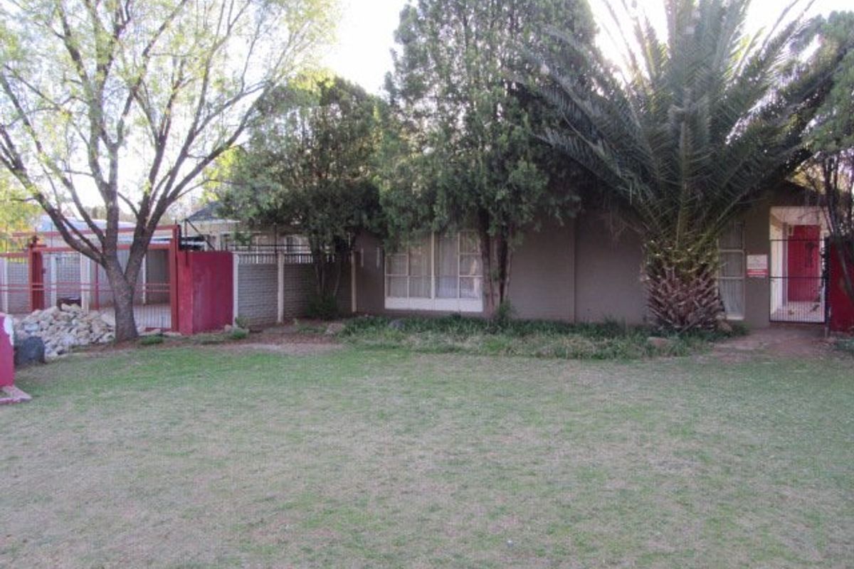 A view of the rear garden featuring a lawn, mature trees, and perimeter security fencing with a metal gate.