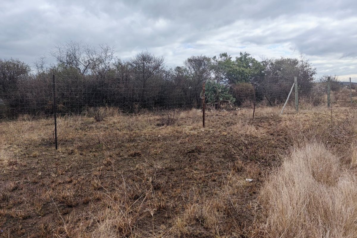 A view of a vacant residential plot featuring dry grass, natural vegetation, and existing wire mesh perimeter fencing.