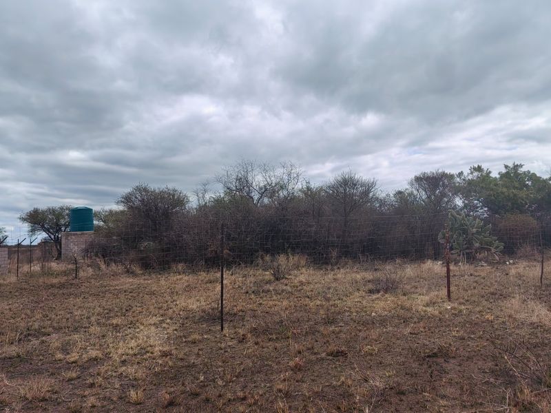A view of a vacant residential plot featuring wire mesh fencing, a water storage tank on a concrete stand, and natural bushveld vegetation.
