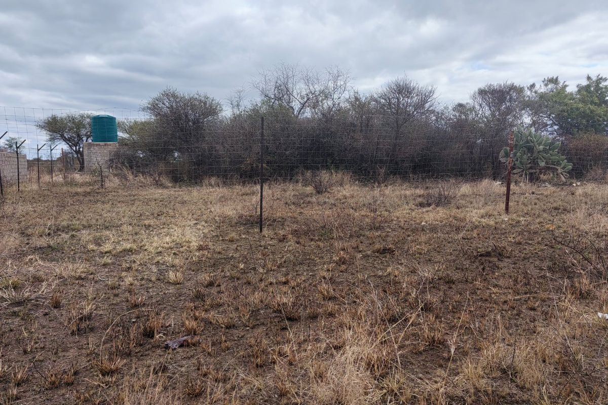 A view of a vacant residential plot featuring dry vegetation, wire mesh perimeter fencing, and a water storage tank on a concrete base.