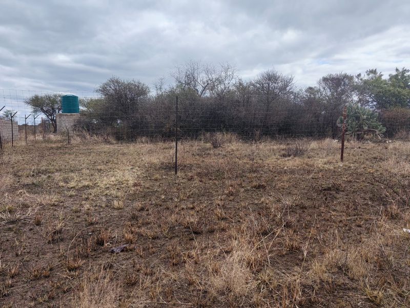 A view of a vacant residential plot featuring dry vegetation, wire mesh perimeter fencing, and a water storage tank on a concrete base.
