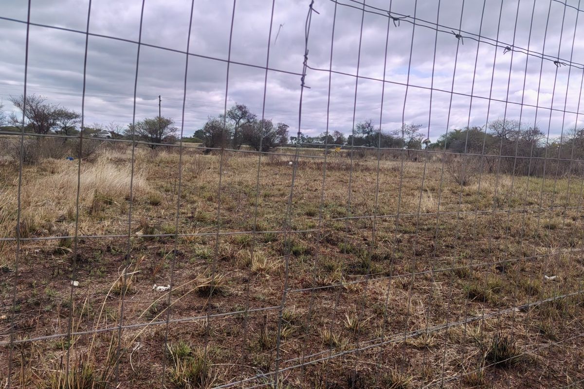 A view of a vacant residential plot enclosed by wire mesh and barbed wire fencing, featuring natural dry grass and scattered shrubs.