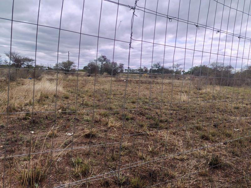 A view of a vacant residential plot enclosed by wire mesh and barbed wire fencing, featuring natural dry grass and scattered shrubs.