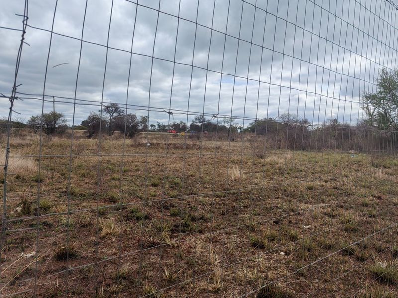 A view of a vacant residential plot enclosed by wire mesh fencing, featuring natural grass and scattered shrubs under an overcast sky.