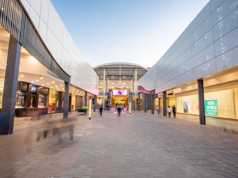 A modern commercial shopping center exterior featuring a wide paved pedestrian walkway, glass storefronts, and a central covered entrance with escalators.