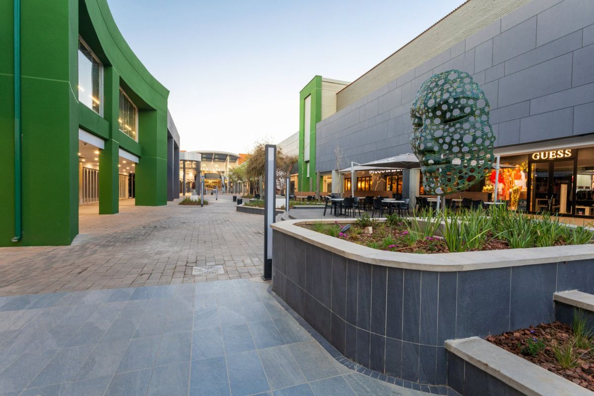 A well-maintained commercial outdoor pedestrian walkway featuring modern tiled facades, landscaped planter boxes, and integrated storefront access points.