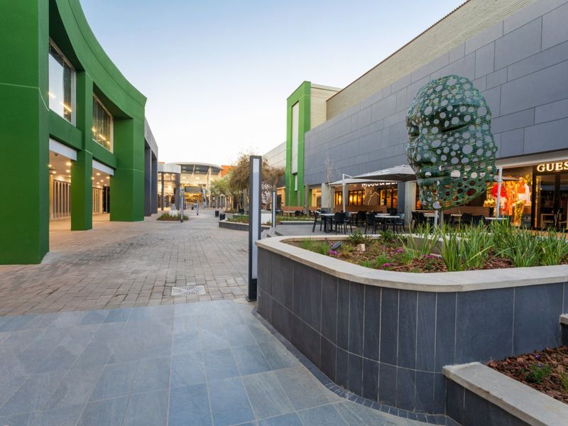 A well-maintained commercial outdoor pedestrian walkway featuring modern tiled facades, landscaped planter boxes, and integrated storefront access points.