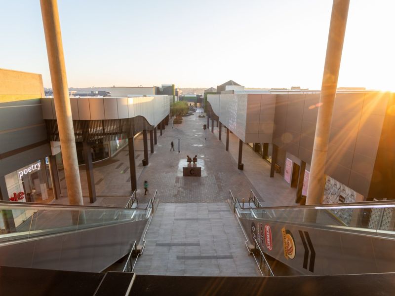 An elevated view of a modern commercial shopping center walkway featuring paved paths, structural columns, and glass storefronts at sunset.