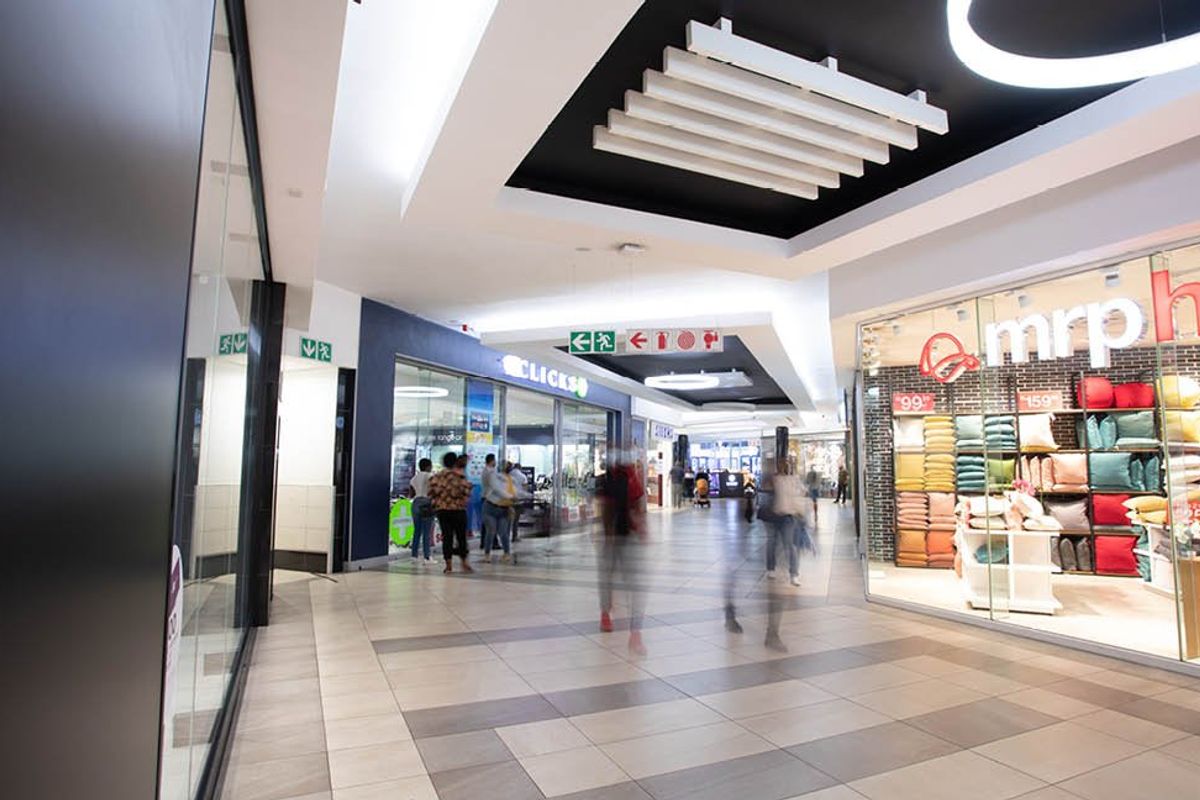 A modern, well-lit commercial shopping centre corridor featuring tiled flooring, glass storefronts, and contemporary suspended ceiling design.