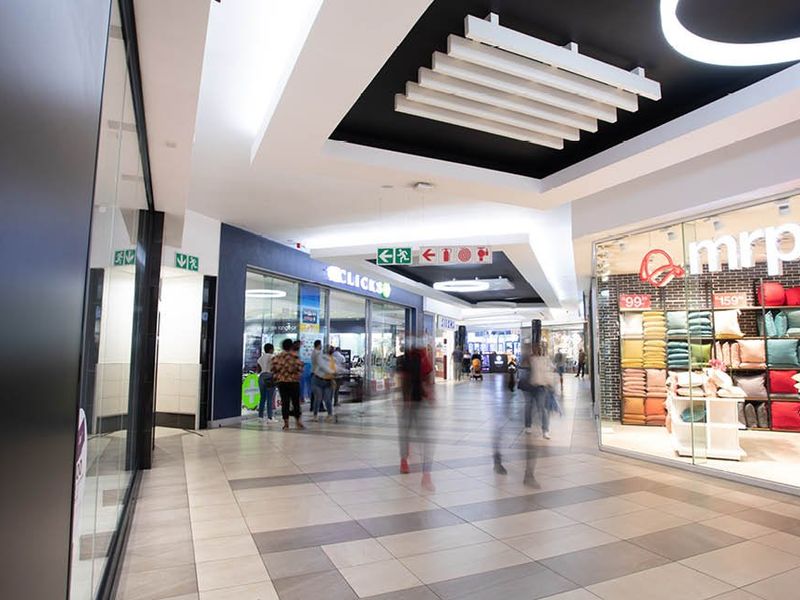 A modern, well-lit commercial shopping centre corridor featuring tiled flooring, glass storefronts, and contemporary suspended ceiling design.