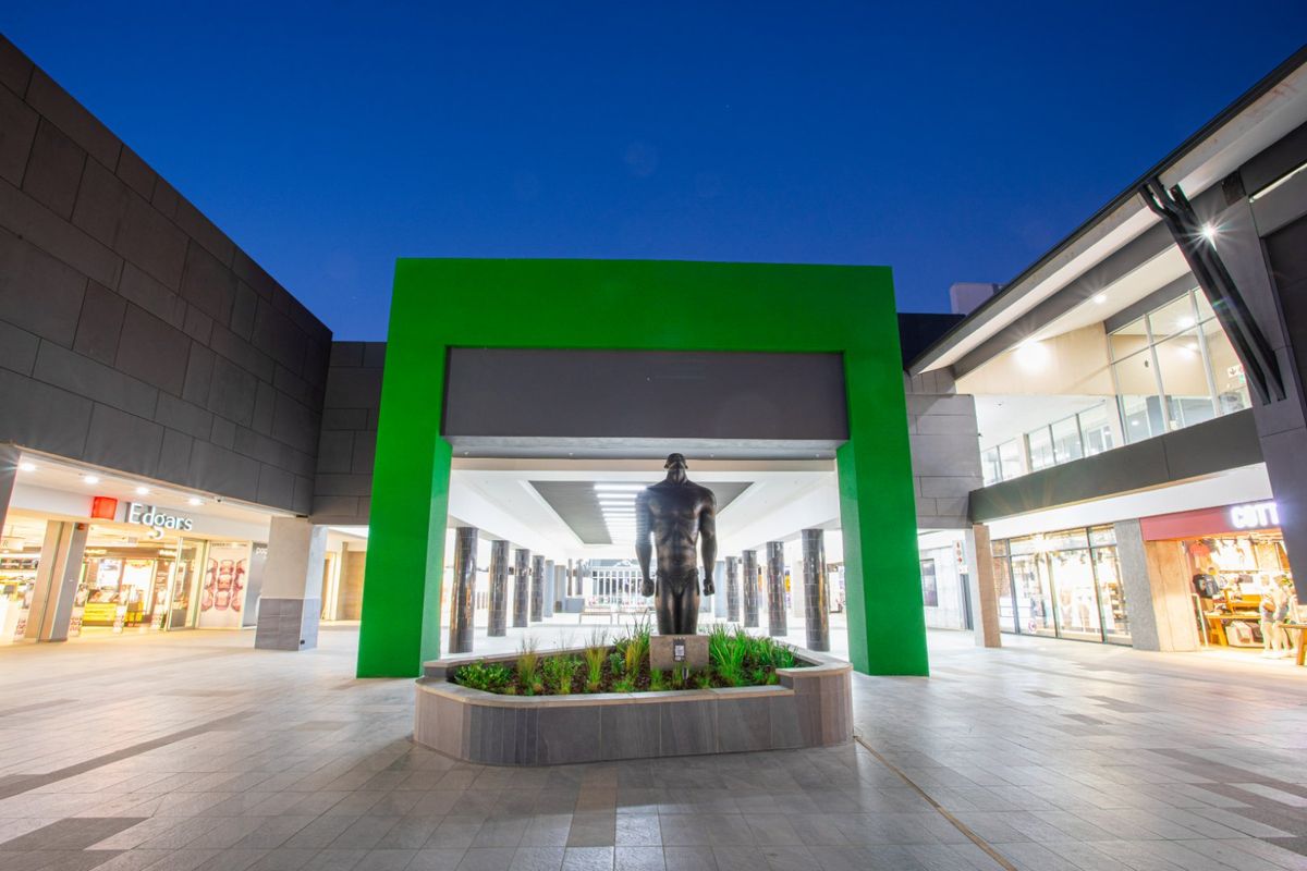 A modern commercial shopping center entrance featuring a central sculpture, paved walkways, and illuminated storefronts under a clear evening sky.