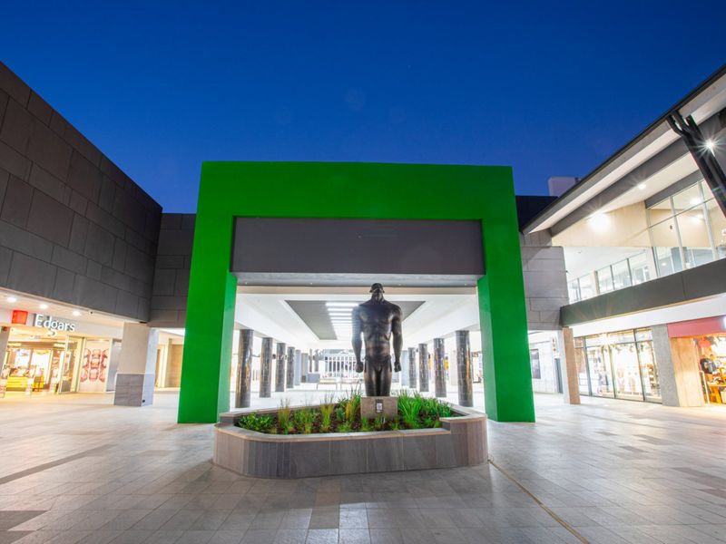 A modern commercial shopping center entrance featuring a central sculpture, paved walkways, and illuminated storefronts under a clear evening sky.
