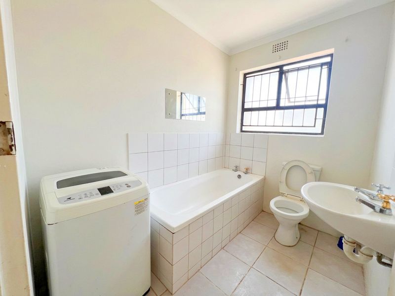 A functional bathroom featuring a tiled bathtub, pedestal basin, and toilet, with security bars on the window and tiled flooring throughout.