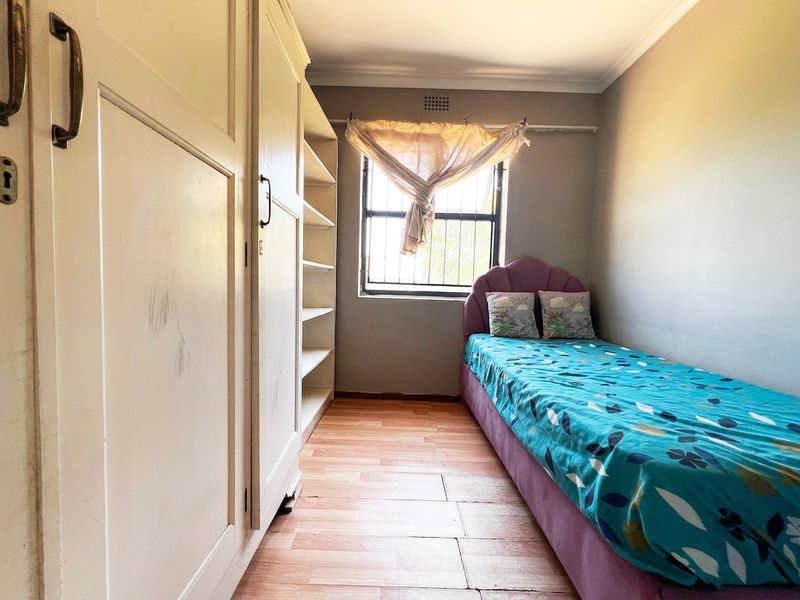 A compact bedroom featuring built-in cupboards, laminate flooring, and a window with security bars, offering a functional space with potential for cosmetic updates.