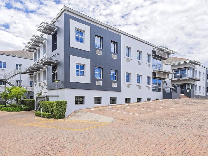 A modern multi-story commercial office building featuring paved parking, external staircases, and contemporary architectural detailing with integrated sunshades and air conditioning units.