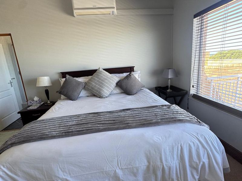 A well-maintained bedroom featuring carpeted flooring, a wall-mounted split-system air conditioner, and large windows fitted with venetian blinds.