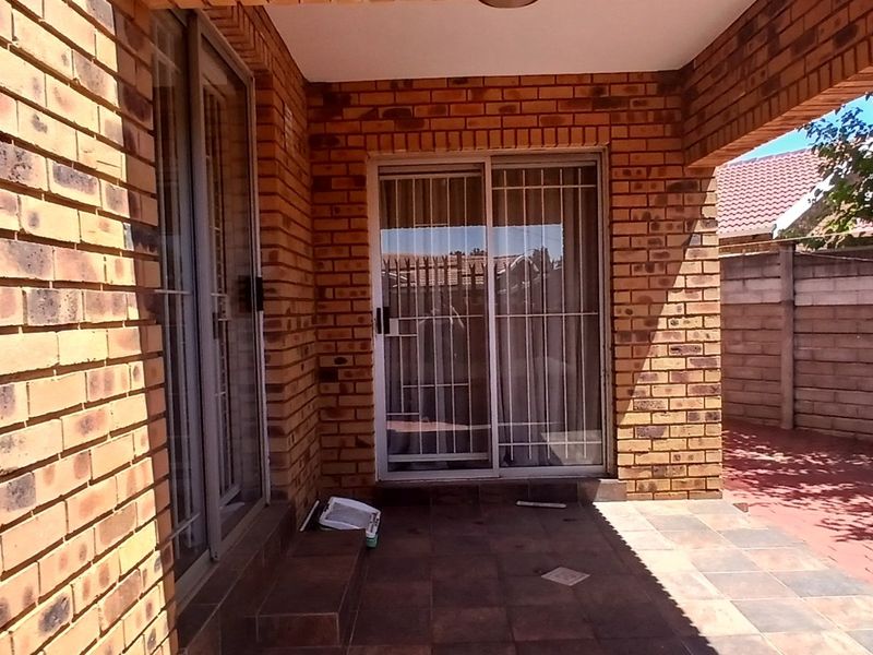 A covered patio area featuring face brick construction, tiled flooring, and sliding glass doors with security gates, providing a functional outdoor transition space.