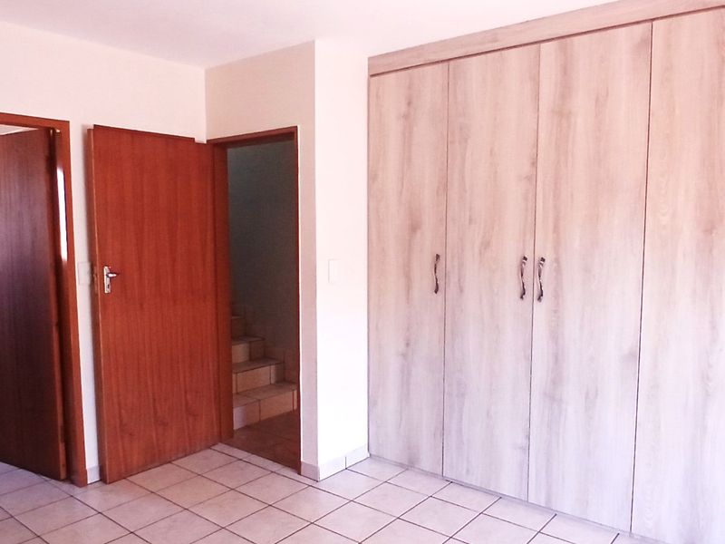 A spacious bedroom featuring light wood-finish built-in cupboards, tiled flooring, and direct access to an en-suite bathroom and hallway.