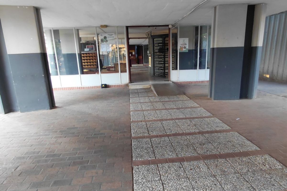 Covered building entrance featuring a paved walkway, access ramp, and security turnstile within a glass-fronted lobby area.
