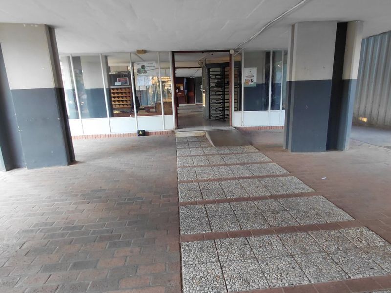 Covered building entrance featuring a paved walkway, access ramp, and security turnstile within a glass-fronted lobby area.