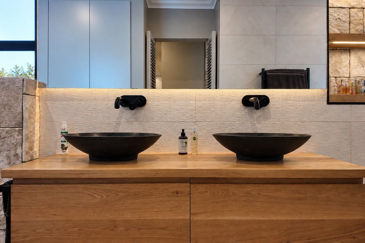 Modern bathroom vanity featuring a floating timber cabinet, dual vessel sinks with wall-mounted matte black faucets, and elegant stone feature wall detailing.