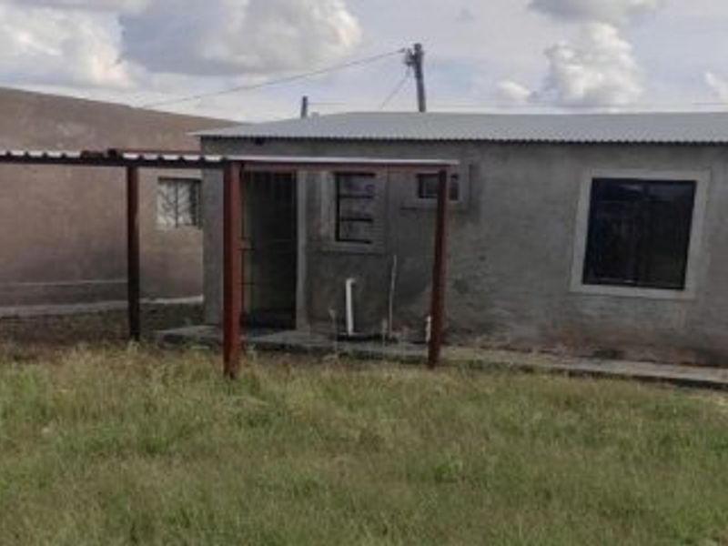 Rear view of a modest residential structure featuring a corrugated metal roof, steel patio frame, and security-barred windows set on a large plot.