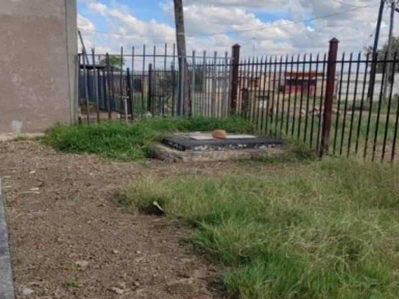 Front yard area featuring metal perimeter fencing, a paved walkway, and an inspection cover set within an unmaintained grass and soil landscape.
