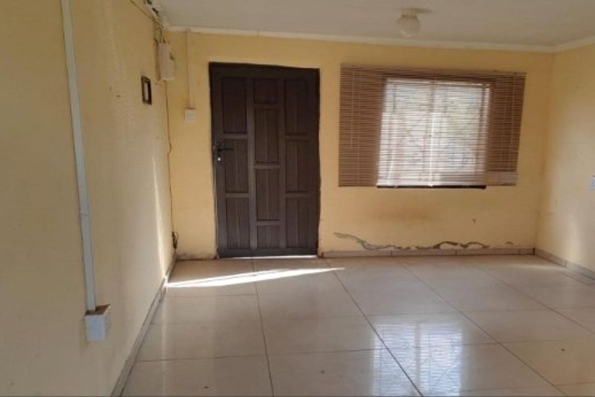 An empty living room featuring tiled flooring, a timber entrance door, and window blinds, showing signs of wall damage and exposed electrical wiring.