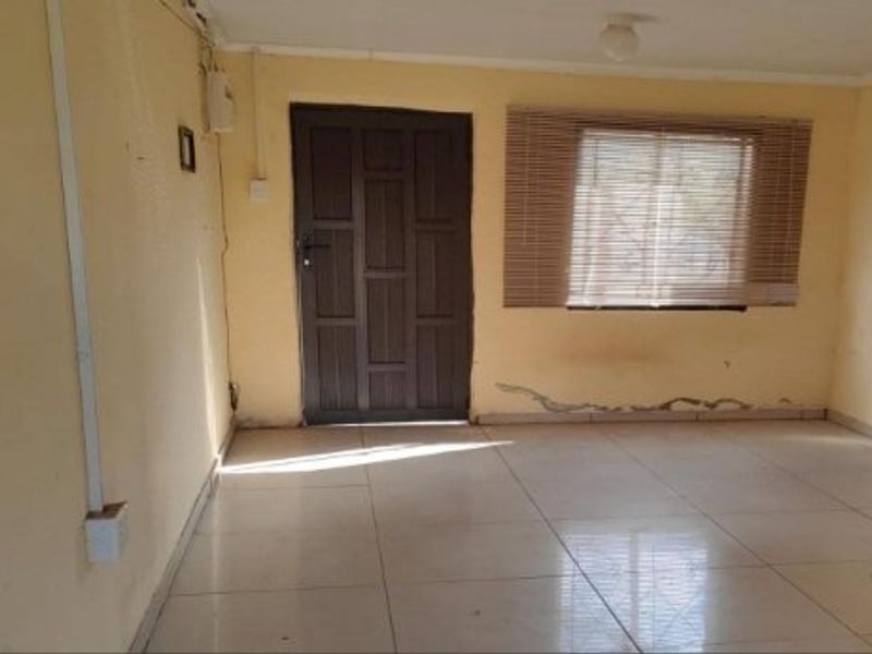 An empty living room featuring tiled flooring, a timber entrance door, and window blinds, showing signs of wall damage and exposed electrical wiring.