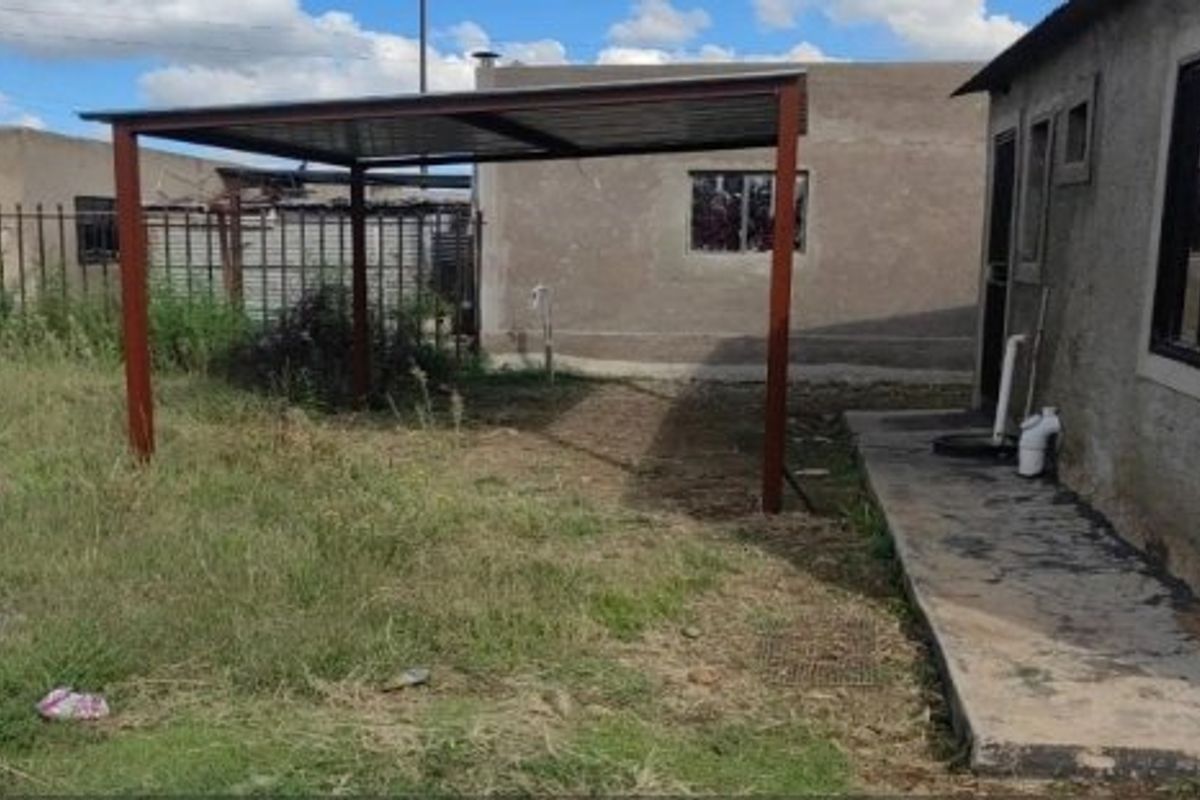 Rear exterior view featuring a metal carport structure, concrete walkway, and steel palisade fencing in a modest backyard setting.