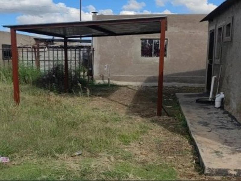 Rear exterior view featuring a metal carport structure, concrete walkway, and steel palisade fencing in a modest backyard setting.