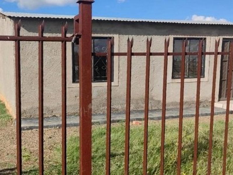 Front view of a single-story house featuring unpainted exterior walls, a corrugated metal roof, and secure metal perimeter fencing.