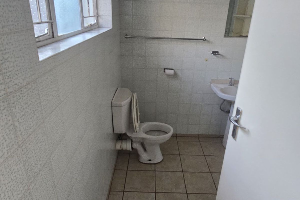 A functional, dated bathroom featuring a pedestal basin, toilet, and full-height wall tiling with neutral floor tiles.