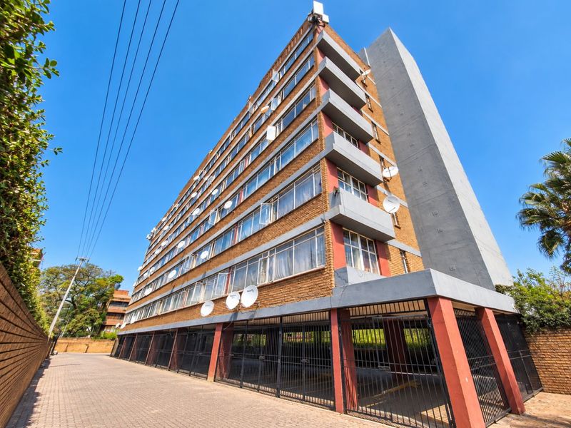 Exterior view of a multi-story apartment building featuring brick facade, private balconies, and a ground-level covered parking area with security gates.