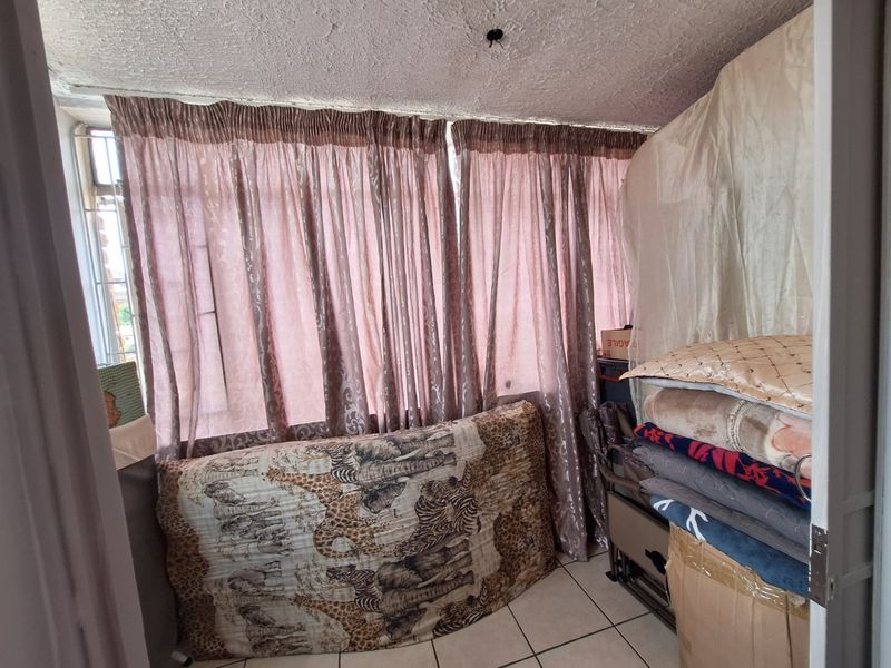 A bedroom featuring tiled flooring and metal security bars on the window, currently used for storage with visible ceiling damage and exposed wiring.