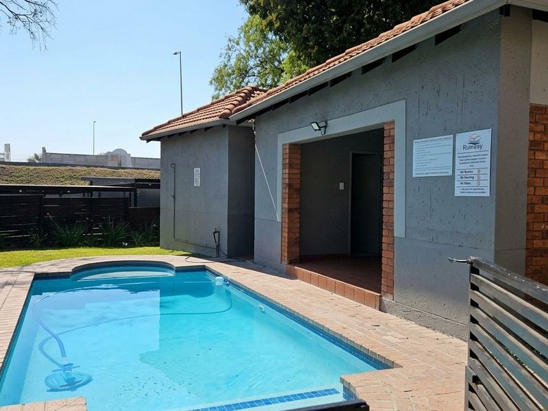 Communal swimming pool area featuring a paved surround, brick-accented building structure, and landscaped garden surroundings.