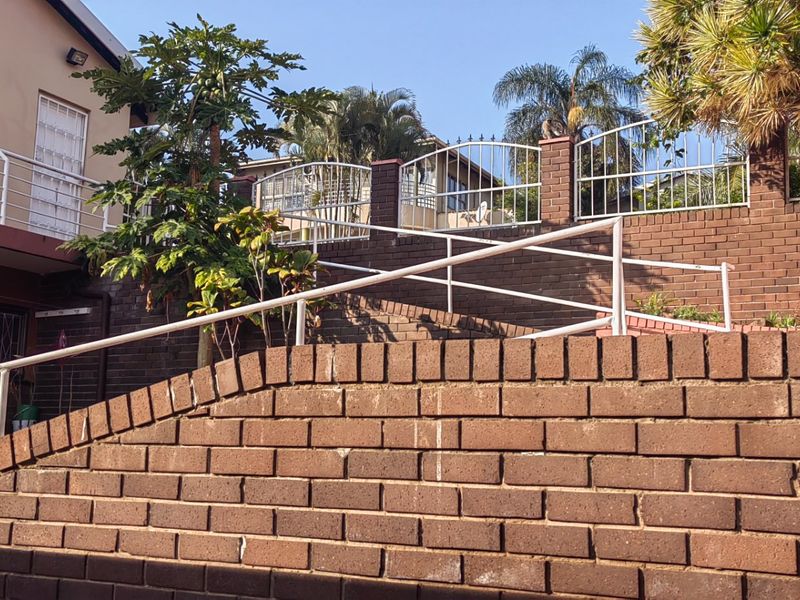 A multi-level outdoor area featuring brick retaining walls, metal handrails, and secure boundary fencing with established garden elements.