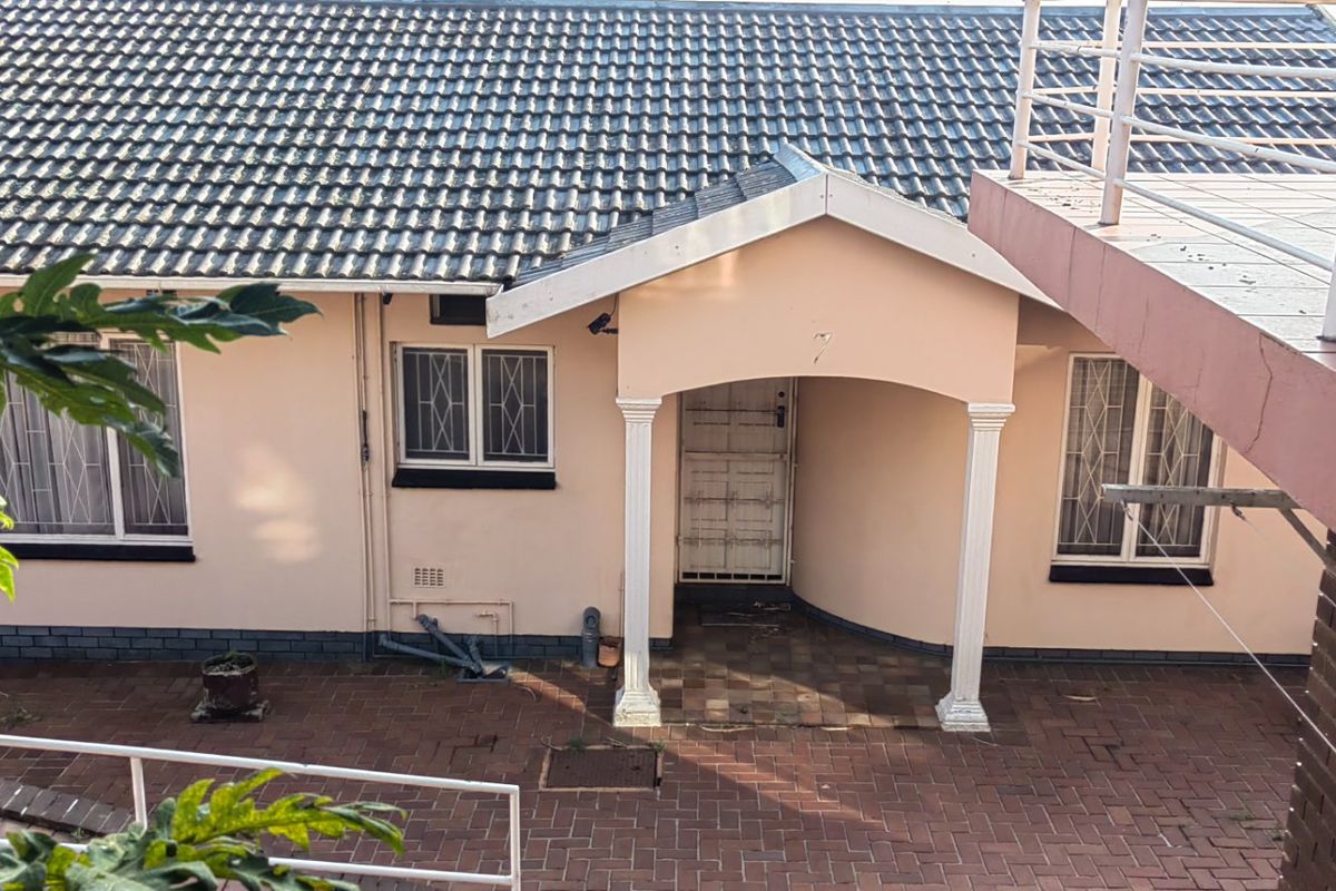 Rear exterior view of the house featuring a paved courtyard, covered entrance with columns, and an upper-level balcony with tiled flooring.