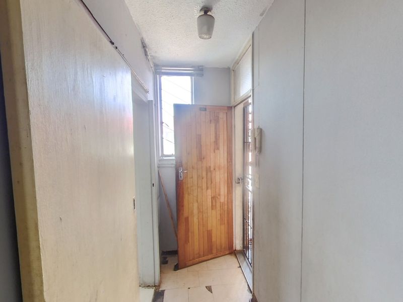 A dated apartment entrance hallway featuring tiled flooring, a timber door, and a security gate, requiring cosmetic updates to improve its overall condition.