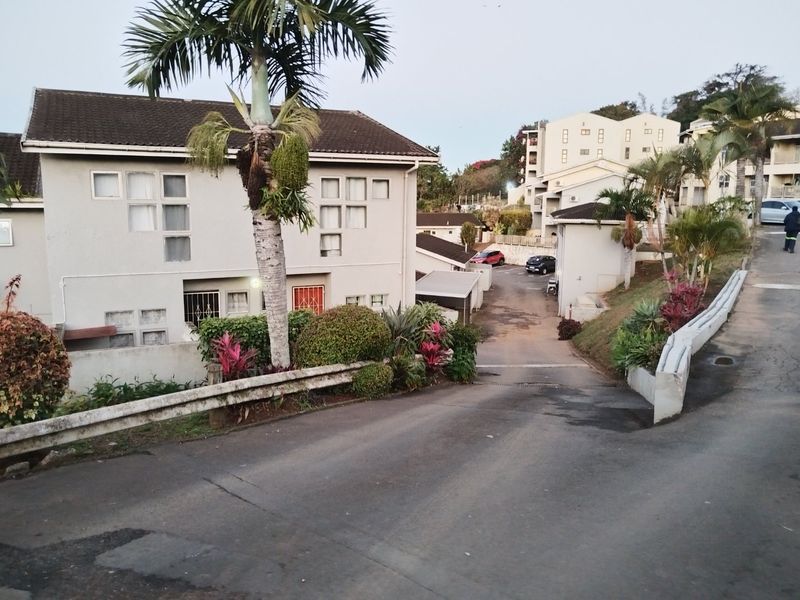 A view of the apartment complex entrance featuring an asphalt driveway, metal guardrails, and established landscaping surrounding the residential buildings.