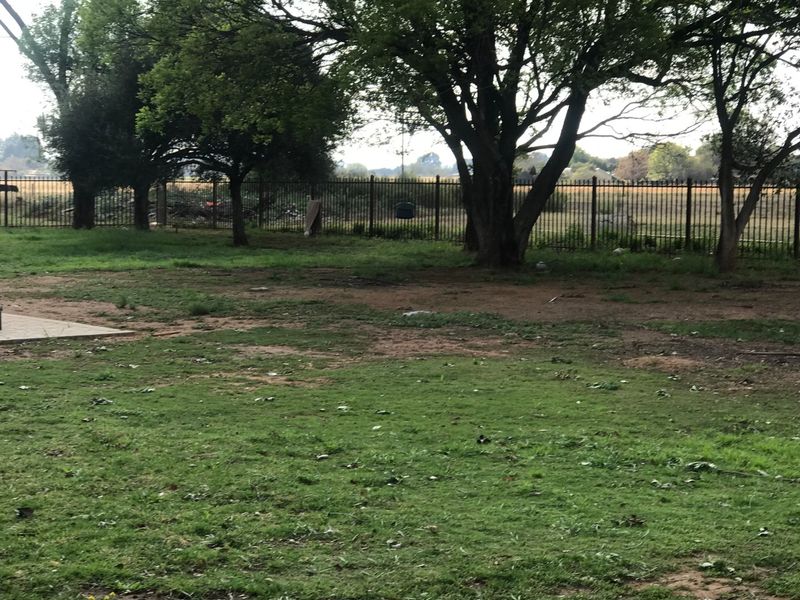 A spacious garden area featuring mature trees and metal perimeter fencing, with areas of patchy lawn and exposed soil.