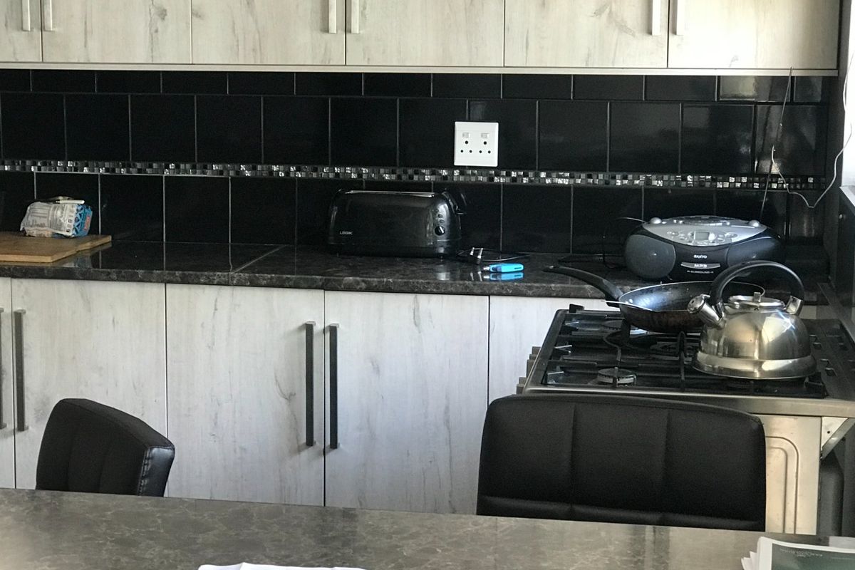 A functional kitchen featuring light wood-grain cabinetry, dark laminate countertops, and a contrasting black tiled backsplash with an integrated gas stove.