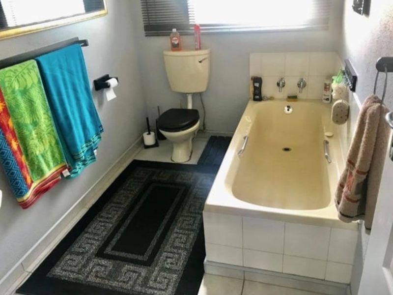 A functional bathroom featuring a built-in bathtub, toilet, and tiled splashback, currently presenting with dated finishes and fixtures.
