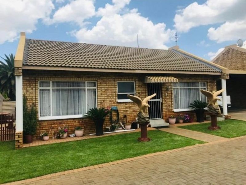 Front elevation of a face brick townhouse featuring a tiled roof, security gate entrance, and a well-maintained front lawn with paved driveway access.