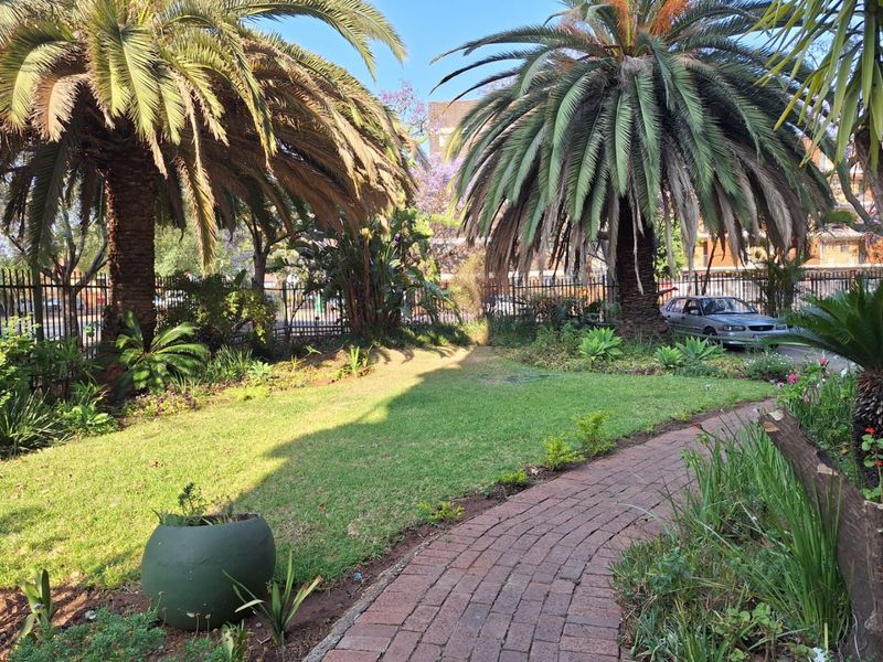 A well-maintained garden area featuring a brick paved pathway, mature palm trees, and secure metal perimeter fencing.
