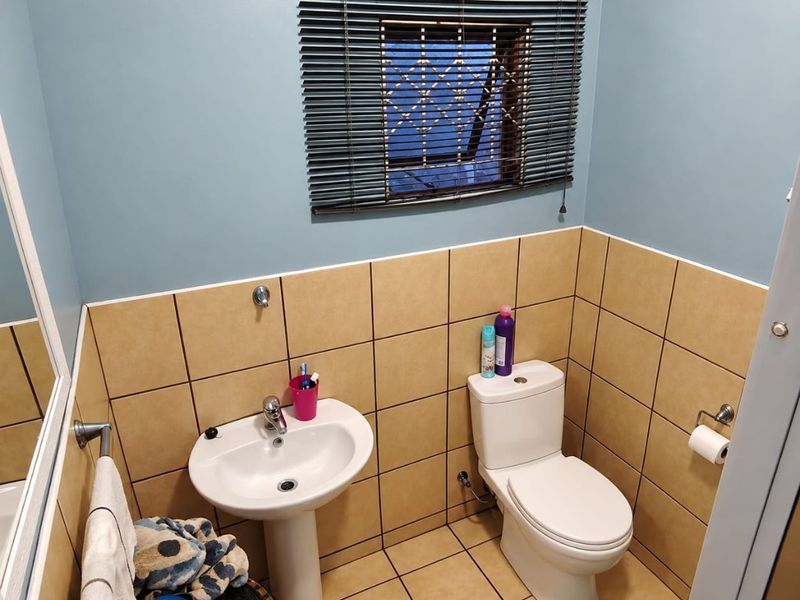 A compact bathroom featuring a pedestal basin, toilet, and neutral wall and floor tiling with a window fitted with venetian blinds.